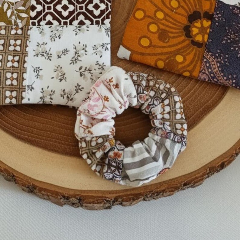 Op de afbeelding: Een patchwork scrunchie met bloemen- en geometrische patronen in wit, bruin en grijstinten. De scrunchie wordt omringd door stoffen vierkanten met vergelijkbare patronen, allemaal op een houten oppervlak.