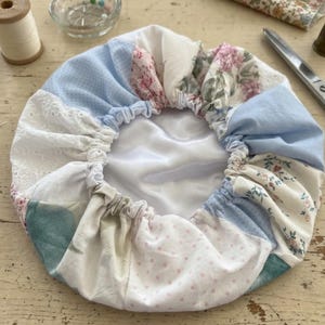 May include: A patchwork bonnet featuring various floral and polka dot patterns in pastel colors. The bonnet has a gathered elastic edge and a white satin lining. The fabrics include blue, pink, and white hues, creating a vintage aesthetic.