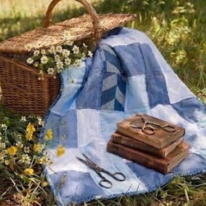 May include: A picnic setting with a wicker basket overflowing with white flowers, a blue and white patchwork quilt, and a stack of old books. A pair of scissors lies on the books and another on the quilt, in a grassy area.