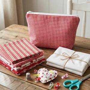 May include: A selection of sewing items on a wooden surface. Features a red and white gingham bag, stacks of red and white patterned fabric, white fabric tied with string, a heart-shaped pin cushion, scissors, and sewing clips.
