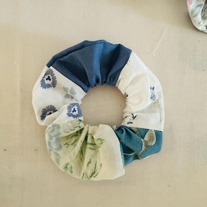May include: A patchwork scrunchie with a mix of floral and solid fabric sections. The scrunchie features a combination of white, blue, and green floral patterns, and solid blue fabric. This hair accessory is a stylish addition to any hairstyle.
