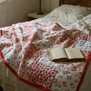 Peut inclure: Une courtepointe faite à la main avec une bordure rouge et un motif de cerises dans des tons de rouge et de rose est posée sur un lit. Un livre ouvert repose sur la courtepointe. Le lit a une jupe de lit en dentelle blanche.