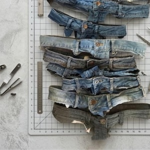 Può includere: Una pila di sette cinture di jeans in denim blu con bottoni in metallo e passanti, disposte su un tappetino da taglio bianco con un righello in metallo visibile. Le tonalità del denim variano dal blu chiaro al blu scuro con texture.
