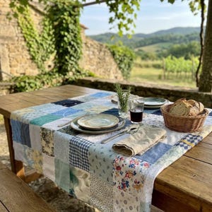 May include: A wooden table set for a meal, featuring a patchwork quilt-style table runner in shades of blue and white. The table is set with plates, silverware, a glass of red wine, and a basket of bread. A sprig of rosemary adds a fresh touch.