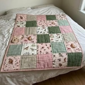 Könnte beinhalten: Eine Patchwork-Decke mit einem sich wiederholenden Raster aus quadratischen Stoffbahnen. Das Design wechselt zwischen einfarbigen Blöcken in Salbeigrün, Altrosa und Beige sowie gemusterten Blöcken mit Illustrationen von kleinen Waldtieren und Pilzen.