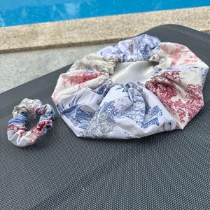 Pode incluir: Uma touca de banho de tecido branco com um padrão floral azul e vermelho. A touca é franzada na parte superior e tem uma faixa elástica branca. Também está incluído um scrunchie a condizer.