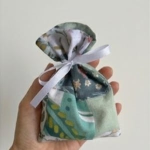 Op de afbeelding: Een kleine patchwork stoffen tas in groen, blauw en wit, vastgebonden met een wit lint. De tas heeft een bloemenpatroon en wordt in een hand gehouden.