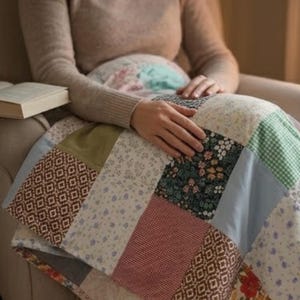 May include: A patchwork quilt featuring a variety of fabric squares in different colours and patterns, including floral prints, gingham checks, and solid olive green. The quilt is draped over a lap, showcasing a mix of cotton-like textile textures.