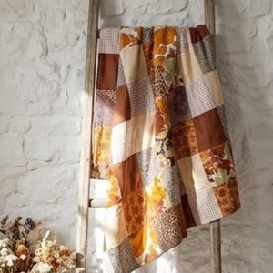 Puede incluir: Un edredón de retazos drapeado sobre una escalera de madera. El edredón presenta cuadrados de varios patrones y colores, incluyendo estampados marrones, beige y florales. El fondo es una pared blanca texturizada.