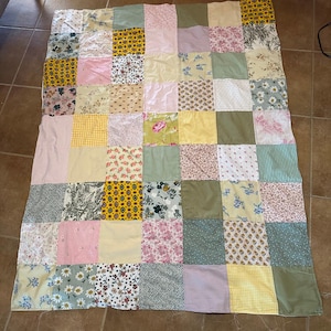 May include: A patchwork quilt with a variety of fabrics in a grid pattern. The quilt features squares of floral, striped, and solid fabrics in shades of pink, yellow, green, and white. The quilt is laid out on a brown surface.