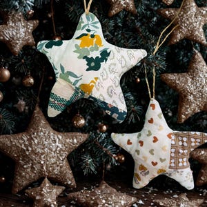 Pode incluir: Dois ornamentos de tecido em forma de estrela pendurados por um cordão. A estrela superior apresenta um design de patchwork com padrões florais amarelos, verde-escuros e brancos. A inferior mostra tecido branco com pequenos corações castanhos.