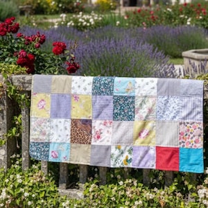 Könnte beinhalten: Ein Patchwork-Quilt mit einer Vielzahl von gemusterten Quadraten, darunter Blumen-, Vichy- und Streifendesigns. Der Quilt ist über einer Steinmauer drapiert, mit einem Blumengarten im Hintergrund. Die Farben des Quilts sind Blau, Gelb, Rot und Grün.