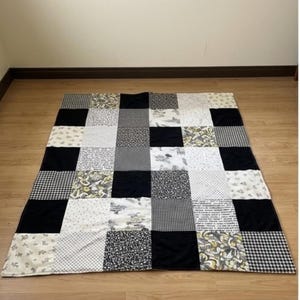 Puede incluir: Una colcha de patchwork rectangular que presenta una cuadrícula de cuadrados de tela alternos en negro, blanco y gris. El diseño incluye cuadrados negros sólidos junto a varios cuadrados estampados con motivos florales, de cuadros, de lunares y abstractos.