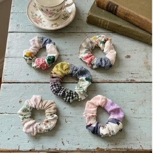 Pode incluir: Cinco elásticos de tecido em várias cores e padrões, incluindo florais, xadrez e tons sólidos. Os elásticos estão dispostos sobre uma superfície de madeira azul clara, com uma chávena e livros no fundo.