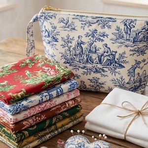 May include: A collection of sewing and crafting supplies on a wooden surface. Includes a toiletry bag with a blue and white pastoral design, stacks of fabric in various colors, a heart-shaped pin cushion, and a thimble.