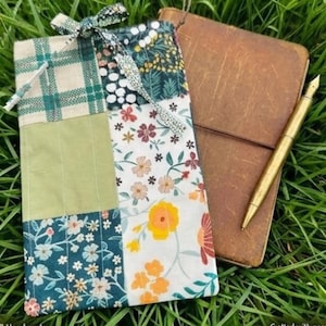 Pode incluir: Uma capa de diário de patchwork com uma variedade de padrões florais e xadrez, amarrada com uma fita. Um diário de couro marrom e uma caneta dourada estão na grama ao lado da capa. Feito à mão.