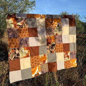 May include: A patchwork quilt in shades of brown, tan, and orange. The quilt is made up of squares with floral, leopard print, and gingham designs. The quilt is displayed on a chain-link fence.