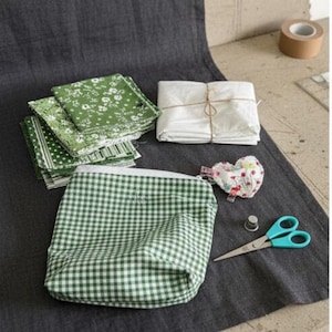 Könnte beinhalten: Eine Auswahl an Nähutensilien auf einer dunkelgrauen Oberfläche. Dazu gehören grün-weiß gemusterte Stoffquadrate, ein grün-weiß kariertes Täschchen, ein herzförmiges Nadelkissen, eine Schere und ein Fingerhut. Eine Rolle Klebeband und ein Bündel weißen Stoffes sind ebenfalls vorhanden.