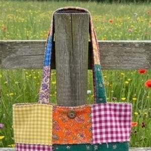 Puede incluir: Un bolso de tela tipo tote hecho de parches con diversos patrones, incluyendo cuadros vichy amarillos, estampado paisley naranja y cuadros morados. El bolso cuenta con dos asas y un botón marrón centrado en el panel frontal.