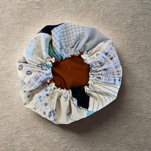 Pode incluir: Um gorro de patchwork com interior castanho e borda elástica. O exterior apresenta vários padrões de tecido em tons de azul, branco e marinho. O gorro foi concebido para proteger o cabelo.