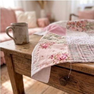 May include: A handmade patchwork quilt with floral and plaid patterns in shades of pink, white, and green. A rustic ceramic mug sits on a wooden table next to the quilt. The quilt is likely used as a decorative throw.