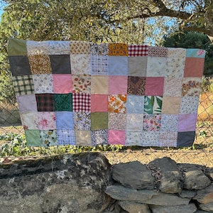 Op de afbeelding: Een patchworkquilt met een verscheidenheid aan stofpatronen en kleuren, waaronder bloemen-, ruit- en effen vierkanten. De quilt wordt buiten tentoongesteld, hangend aan een draadomheining, met een stenen muur op de voorgrond.