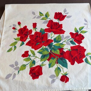 May include: A white fabric tea towel featuring a central floral print of vibrant red roses with green leaves and light gray foliage accents. The design displays multiple blooming rose heads and buds arranged in a cluster across the center.