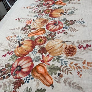 May include: A rectangular tablecloth with a light beige linen-like texture featuring a central autumn-themed print. The design includes orange and red pumpkins, gourds, brown acorns, red berries, and colorful fall leaves with floral accents.