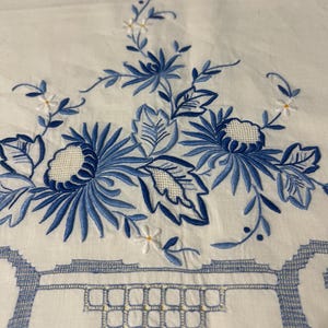 May include: White fabric with intricate blue floral embroidery. The design features stylized blue flowers, leaves, and delicate white accents. The bottom edge has a geometric border, creating a classic, elegant look. This textile is suitable for home decor.