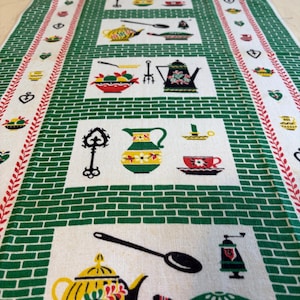 May include: A rectangular kitchen towel featuring a white background with a green brick pattern border. The center displays repeating rectangular panels containing illustrations of kitchenware including yellow teapots, red cups, green pitchers, spoons, and keys.