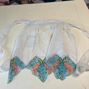 May include: A white apron with a floral design. The apron has a pocket and ties at the waist. The bottom edge features a scalloped design with pink and blue flowers and green leaves.