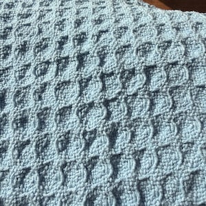 May include: A light blue crocheted blanket featuring a repeating waffle stitch pattern. The texture consists of raised square grids created by interconnected yarn loops, resulting in a consistent, dense, and tactile surface throughout the fabric.
