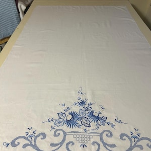 May include: White tablecloth with a detailed blue floral and scrollwork embroidery design. The embroidered section is located at the bottom of the tablecloth. The design includes flowers, leaves, and decorative scroll patterns.