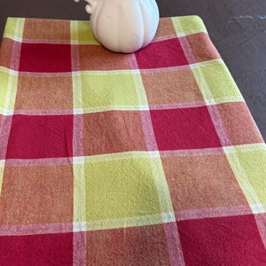 May include: A white ceramic pitcher with a curved handle and a fluted body sits atop a rectangular fabric cloth featuring a large plaid pattern in shades of deep red, lime green, and orange, separated by thin white intersecting grid lines.