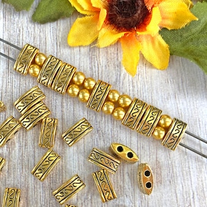 May include: A collection of gold-toned metal beads with a decorative swirl pattern. The beads are arranged on a wooden surface with two metal beading needles.