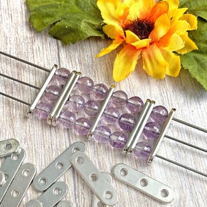 May include: A close-up of a silver bracelet with lavender beads. The bracelet is made with three silver bars and has a simple design. There are also several silver connector pieces scattered around the bracelet.