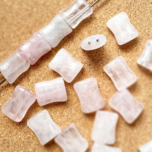May include: Rose quartz and clear quartz beads in various shapes. Some are strung on a wire, while others are loose. A small, white, oval-shaped bead with two holes is also present. The beads are on a corkboard surface.