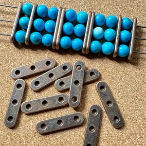 May include: A close-up shot of a handmade bead component. The component features bright turquoise beads strung on silver wire, with several small, rectangular metal pieces with three holes each. The background is a light brown, textured surface.