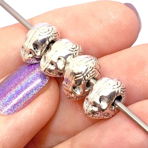 May include: A close-up of five silver skull-shaped beads strung on a wire. The beads have intricate details and a slightly aged finish.