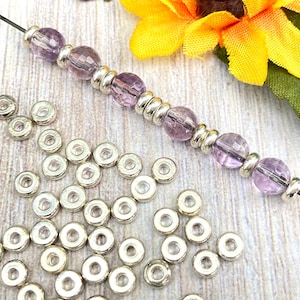 May include: A close-up of a strand of purple beads with silver spacer beads. Scattered around the beads are many silver spacer beads.