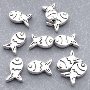 May include: A collection of silver-toned metal fish-shaped beads. Each bead features a stamped, dark-accented design depicting a fish with a circular eye, a curved gill line, and a zigzag pattern along the body. The beads are arranged in a scattered layout.