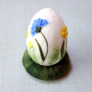 Op de afbeelding: Een wit vilten ei versierd met blauwe en gele bloemen en groene bladeren. Het ei zit op een groene viltbasis.
