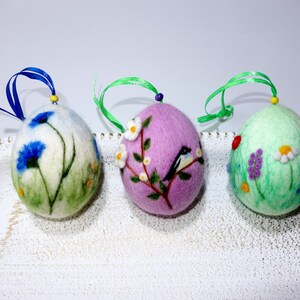 Op de afbeelding: Drie vilten paaseieren met verschillende lentethema's. Eén ei is groen met paarse bloemen, een ander is roze met een vogel en witte bloemen, en de laatste is wit met blauwe bloemen.