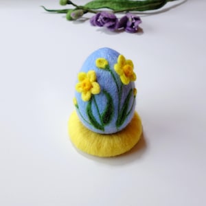 Op de afbeelding: Een blauw vilten paasei versierd met gele narcissen en groene bladeren. Het ei ligt op een gele viltbasis.