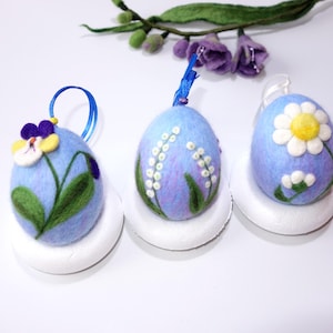 Op de afbeelding: Drie blauwe vilten paaseieren versierd met verschillende lentebloemen. De eieren staan op witte schuimrubberen bases.