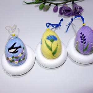 Op de afbeelding: Drie vilten paaseieren versierd met geborduurde bloemen en een vogel. De eieren worden gepresenteerd op witte schuimrubberen bases.