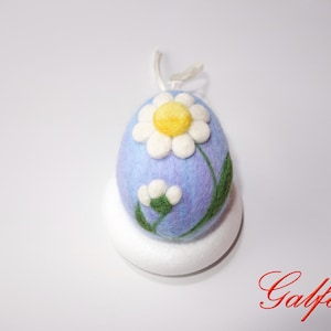 Op de afbeelding: Een blauw vilt paasei met een witte madeliefje en groene bladeren. Het ei staat op een witte voet.