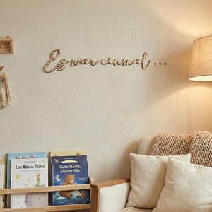 May include: Wooden wall decor with the phrase "Es war einmal..." in cursive script. The room features a bookshelf with children's books, a cozy armchair with pillows, and a woven blanket. A lamp provides soft lighting.
