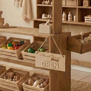 Peut inclure: Étalage de magasin de jouets en bois avec des caisses remplies de fruits, de légumes et d'autres jouets en bois. Une enseigne indique "Nila and Hanna's shop OPEN". Le magasin a des étagères avec des voitures jouets, des bouteilles et d'autres jouets en bois.