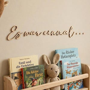 May include: Wooden bookshelf with children's books and a crocheted bunny. The books include "Emil und die Detektive", "Die kleine Hexe", "Der R&auml;uber Hotzenplotz", and "Das fliegende Klassenzimmer". Above the shelf is wooden text that reads "Es war einmal..."